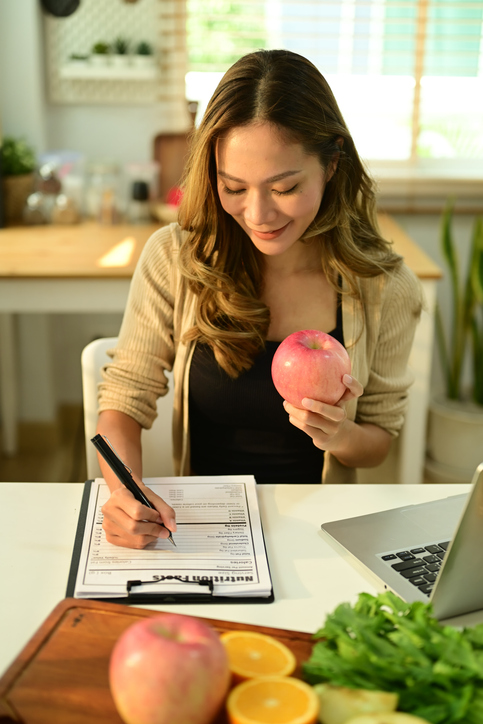 Charming young woman holding an apple and writing recipe at desk with fresh fruit.