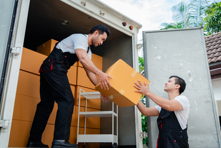 two men movers in uniform lifting boxes unloading boxes and furniture from a moving truck.