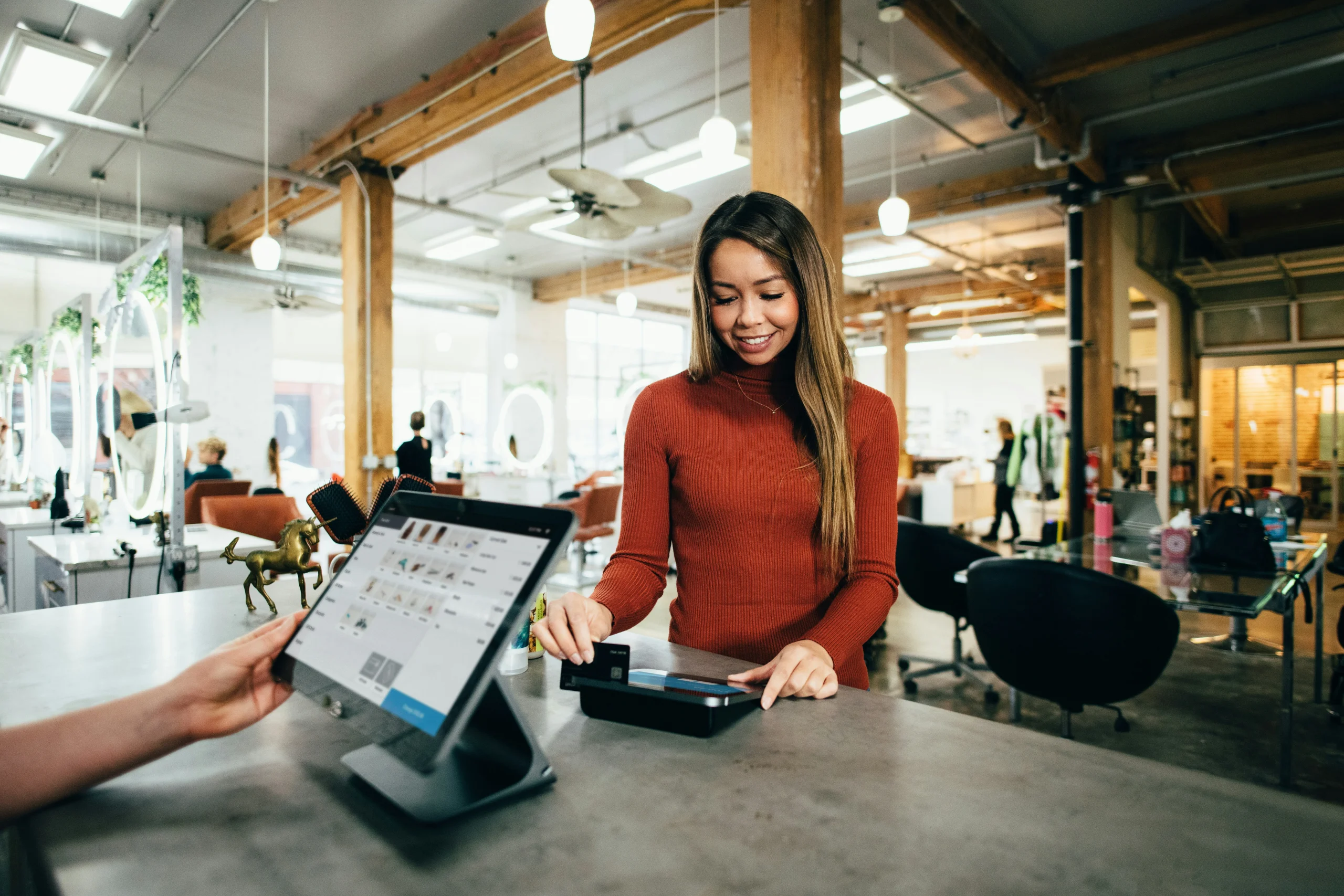 A woman in a red sweater uses her credit card to check out at a payment terminal while a clerk helps from behind a computer screen.