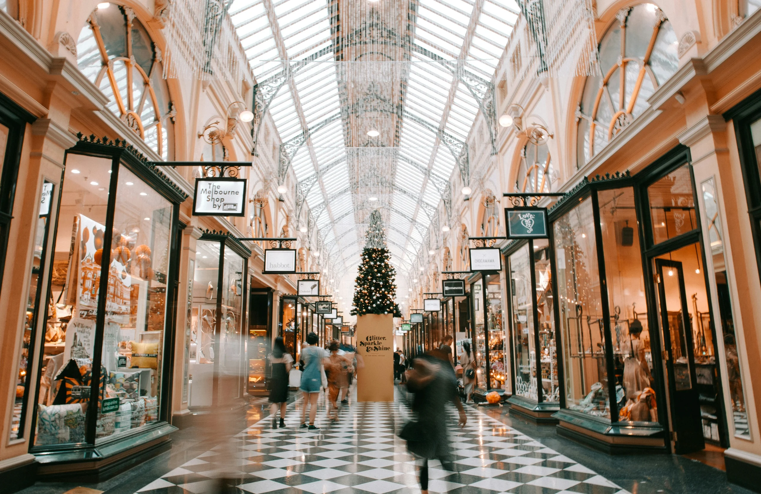 Image of an in door retail space. Each store has a glass store front. Above is a domed glass ceiling letting in natural light. A Christmas tree stands at the far end of the corridor.