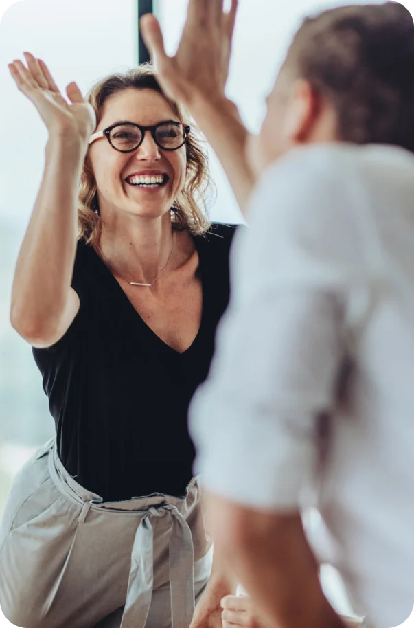 Smiling woman wearing glasses gives a high-five to another person in a bright indoor setting.